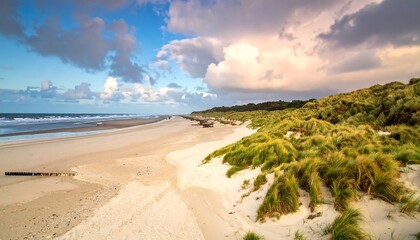 Panoramic view of a serene beach at sunset, with sand dunes covered in lush green grasses and a partly cloudy sky