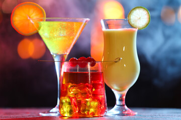 Glasses of alcoholic cocktails with garnish on table against blurred background with smoke, bokeh effect
