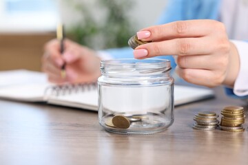Woman putting coins into glass jar while working at wooden table indoors, closeup