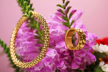 Stylish gold ring, bracelet and beautiful flowers against pink background, closeup
