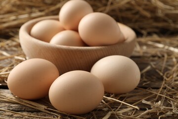 Raw chicken eggs in bowl and straw on wooden table, closeup