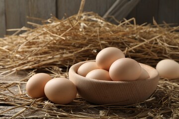 Raw chicken eggs in bowl and straw on wooden table, closeup