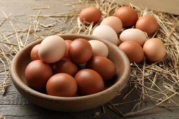 Raw chicken eggs in bowl and straw on wooden table, closeup