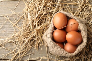 Raw chicken eggs in burlap sack and straw on wooden table, top view. Space for text
