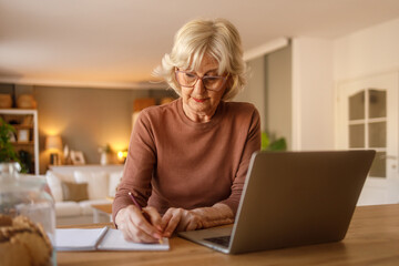 Focused senior woman in eyeglasses writing notes while paying utilities online through laptop on desk at home
