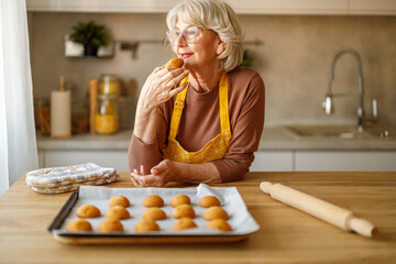Happy senior woman in eyeglasses looking away and eating cookie while confidently sharing recipe on her blog