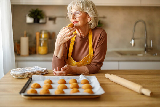 Happy senior woman in eyeglasses looking away and eating cookie while confidently sharing recipe on her blog