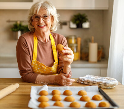 Portrait of happy senior woman holding cookie while leaning by tray of freshly baked sweets on kitchen island at home