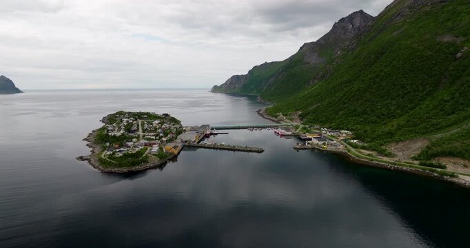 Hus&oslash;y fishing village on island in &Oslash;yfjorden, harbor, bridge, and majestic mountains, Senja, Norway. Aerial backward, copy space