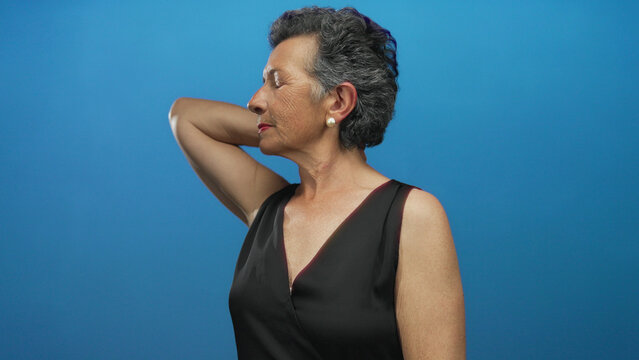 Senior woman with grey hair in black dress against blue background, expressing neck pain, with closed eyes and relaxed posture, conveying discomfort yet calm demeanor.