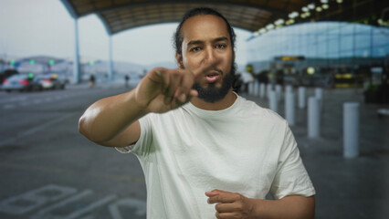 Fototapeta premium Man points finger at camera at airport terminal curbside, wearing white t shirt with beard and clasped hands; confidence.