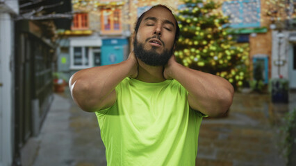 Obraz premium Man wearing bright green shirt holding neck with both hands on wet cobblestone street, grimacing from neck pain; pain recovery.