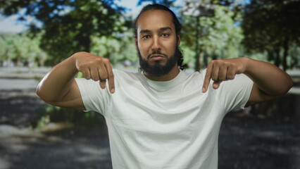 Naklejka premium Man with beard in white t shirt points both index fingers down toward his shirt in forest setting, direct stare at camera; confidence.