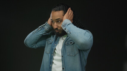 Man wearing denim jacket with hands covering ears and pained expression on black studio backdrop;...