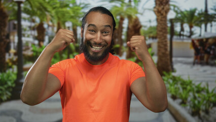 Man cheering with clenched fists and a wide smile on street lined with palm trees in a green park; joy celebration.