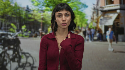Hispanic woman in a red cardigan pointing finger to herself on a busy city street with parked...