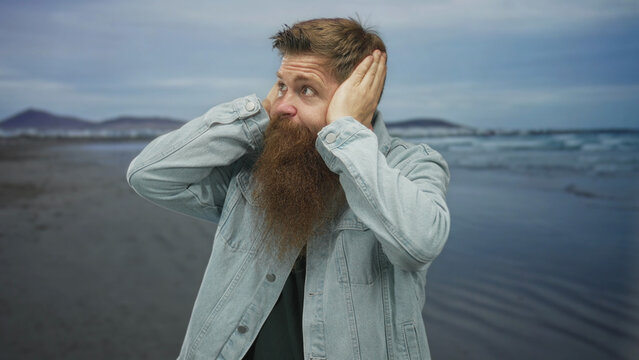 Bearded young man covers ears with both hands at windswept beach shore; overwhelming noise frustration.