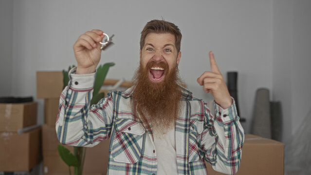 Man with beard holds house key and grins amid stacked moving boxes inside new building foyer; excitement.