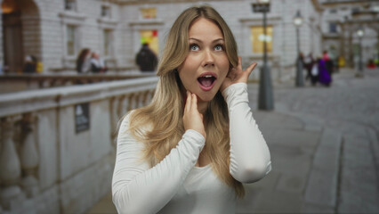 Fototapeta premium Blonde woman with hand on cheek and mouth open on a busy city sidewalk in daylight; surprise awe amazement excitement.