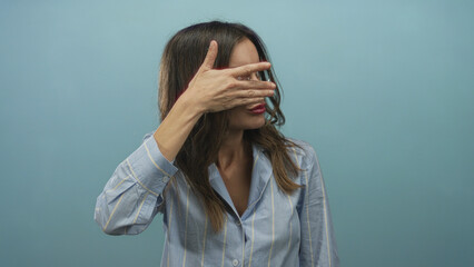 Woman covering her eyes with her hand against a blue studio backdrop while wearing a striped shirt; mystery.