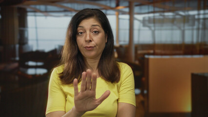 Woman wearing yellow shirt shows hand palm to camera in hotel interior with serious look and...