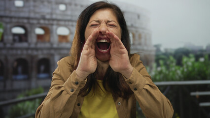Woman in brown jacket cups hands around mouth to shout at roman coliseum building facade;...