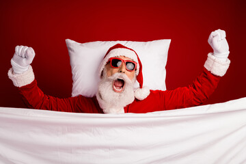 Cheerful Santa Claus wearing sunglass waking up in festive spirit on a cozy bed with white bedding...