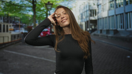 Fototapeta premium Woman with hand to hair on street by canal and building, smiling with visible face and bare neck; carefree joy.