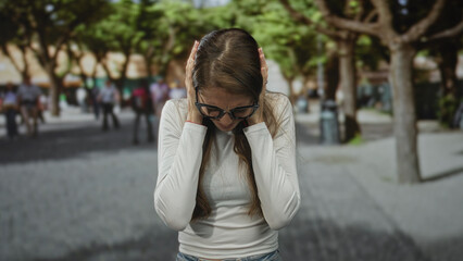 Woman covering ears and holding head, wearing glasses and a white top on a city street lined with trees and pedestrians, reacting to noise and commotion; urban frustration.