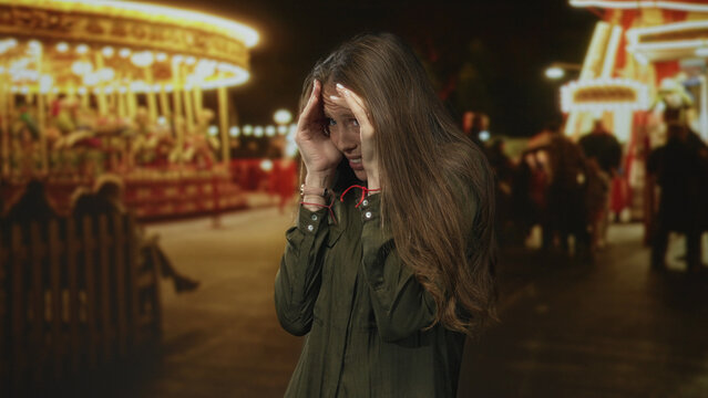 Young hispanic woman covering face with hands at crowded street fair by lit carousel and fence at night; anxiety.