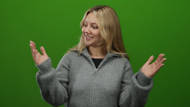Woman smiling joyfully against an isolated green background with blonde hair, wearing a grey sweater, gesturing with hands in a cheerful manner.
