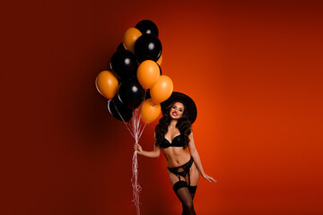 Witch in glamorous lingerie holds festive balloons on orange background celebrating Halloween joyfully