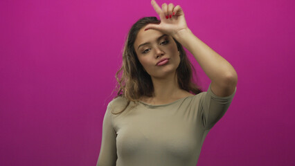Hispanic woman wearing beige top makes loser gesture with hand and raised brow in pink studio;...