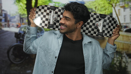 Man in a denim shirt smiling holds a checkered skateboard behind his head under sunlit trees on a...