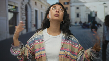 Fototapeta premium Woman touches hair with one hand and shrugs on a cobblestone street by buildings, wearing glasses and patterned jacket; exasperation routine.