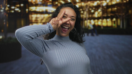 Woman smiling with visible teeth and flashing peace sign near eye on street at night; joyful playfulness.