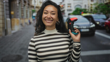 Woman holding a game controller and points finger to controller in city street crosswalk smiling broadly  gaming playful joy. © Krakenimages.com