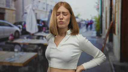 Young blonde woman holds her lower back with a pained expression at an outdoor restaurant terrace...