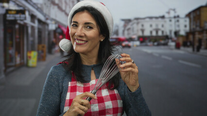 Middle aged hispanic woman holding whisk on city street smiling in red checkered apron and santa...