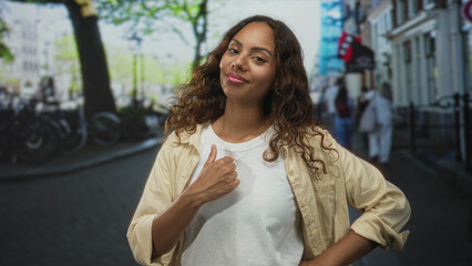 Fototapeta premium Woman points thumb to chest in casual pose on a city street with bicycles and row buildings visible; pride self belief.