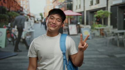 Young man smiling and making a peace sign on an outdoor terrace of a coffee shop with a relaxed atmosphere in a bustling urban restaurant setting.