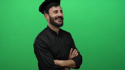 Smiling hispanic male chef with beard in black uniform standing confidently against vibrant green background showcasing culinary expertise.