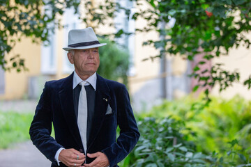 An elderly man in official clothes confidently walks along an open path surrounded by herbs