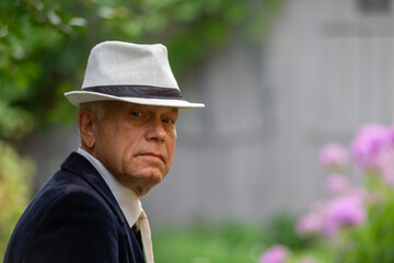 Older man wearing a stylish hat looks thoughtfully at the camera in a vibrant garden setting during a sunny day