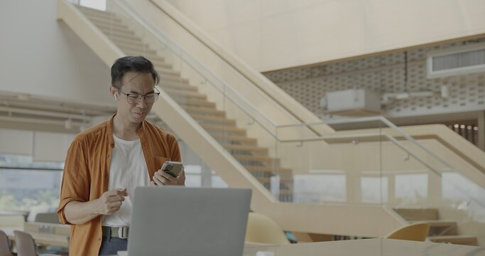Senior Asian businessman wearing headset celebrates success during online meeting in modern co-working space. Remote work and business communication concept