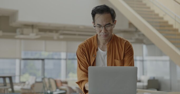 Senior Asian businessman wearing headset celebrates success during online meeting in modern co-working space. Remote work and business communication concept
