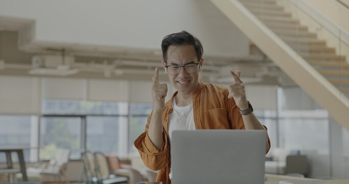 Senior Asian businessman wearing headset celebrates success during online meeting in modern co-working space. Remote work and business communication concept