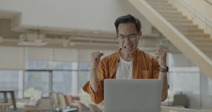 Senior Asian businessman wearing headset celebrates success during online meeting in modern co-working space. Remote work and business communication concept