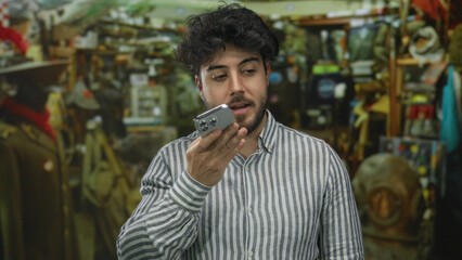 Young hispanic man with beard recording voice message on smartphone in indoor bazar surrounded by...