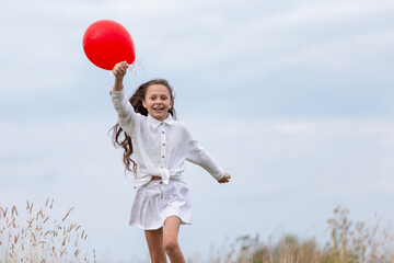 Young girl runs joyfully in a field holding a bright red balloon on a cloudy day